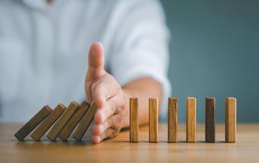 A man's hand preventing a cascade of wooden blocks, demonstrating control or intervention in a chain reaction, with a plain background