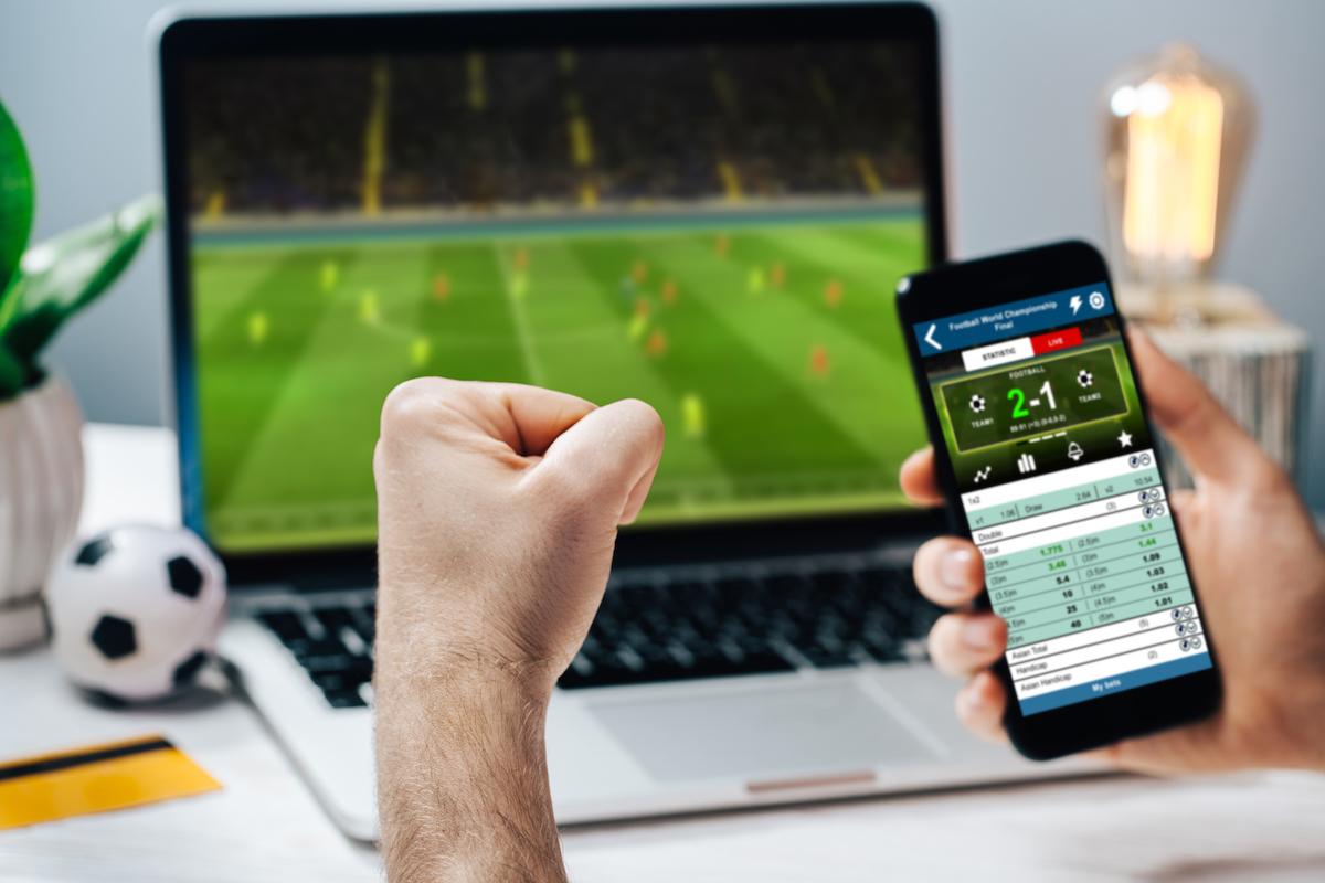 An excited fan watches a football match on a laptop while simultaneously checking live scores and placing bets on a smartphone
