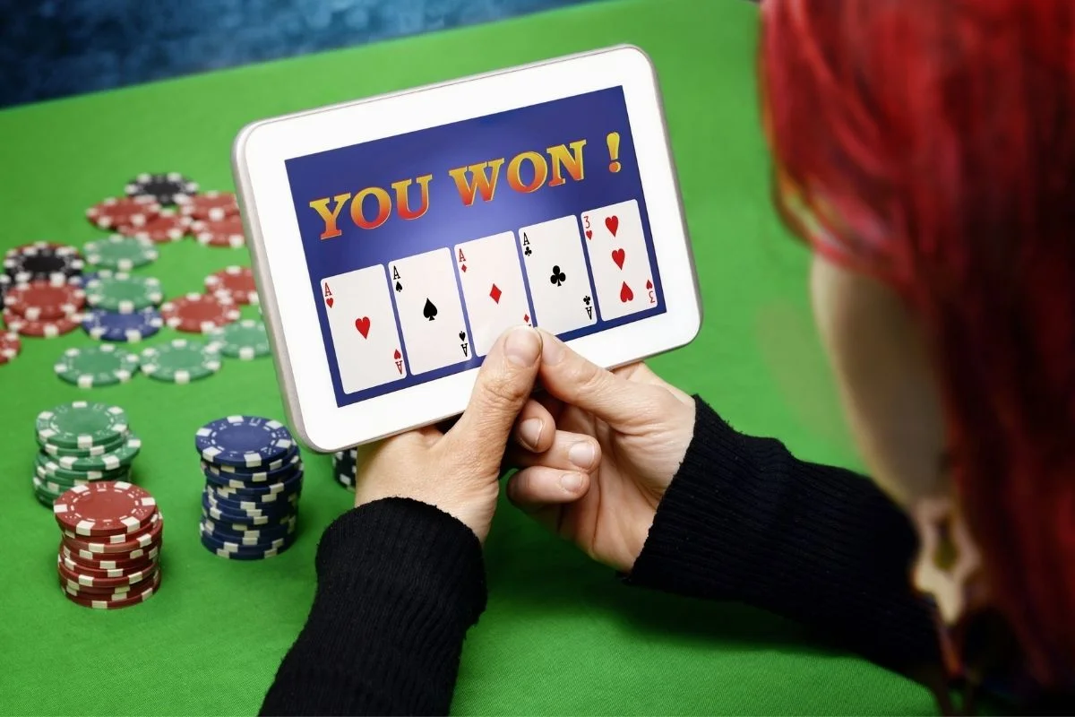 A young woman's hands hold a tablet displaying "YOU WON!" and a five-card poker hand of four aces and a king, with stacks of casino chips on a green felt table in the background